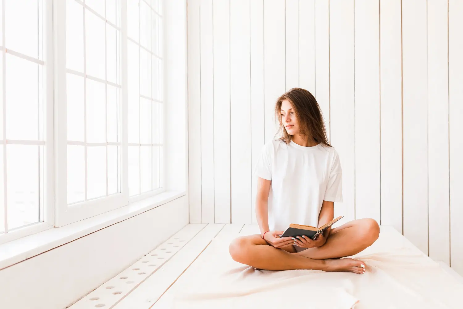 A young woman sitting cross-legged on a bed by a bright window, holding an open book