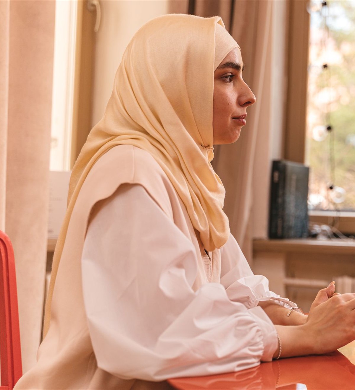 A woman in a cream hijab sitting at a table by a window in warm daylight, in conversation