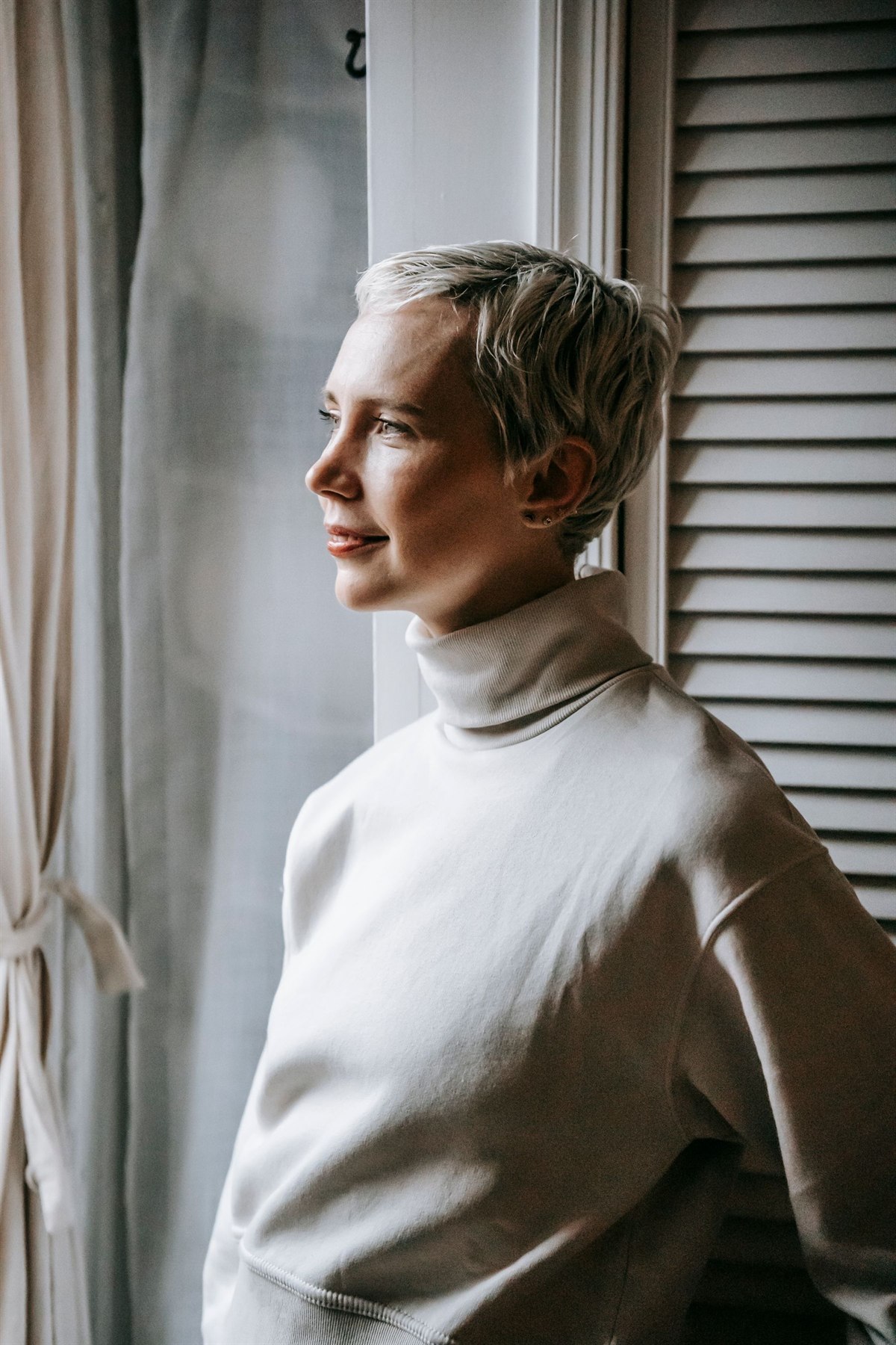 A woman in a cream turtleneck standing by a window in soft daylight, looking out