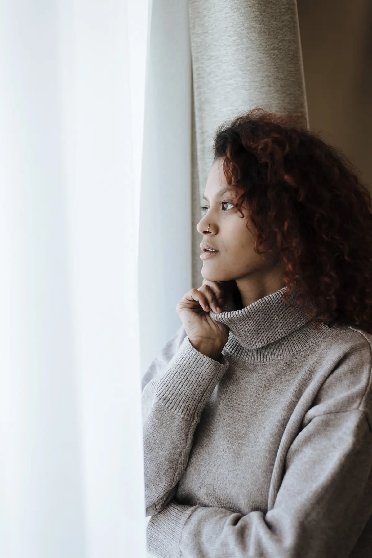 A woman in a warm sweater looking out of a window, thinking