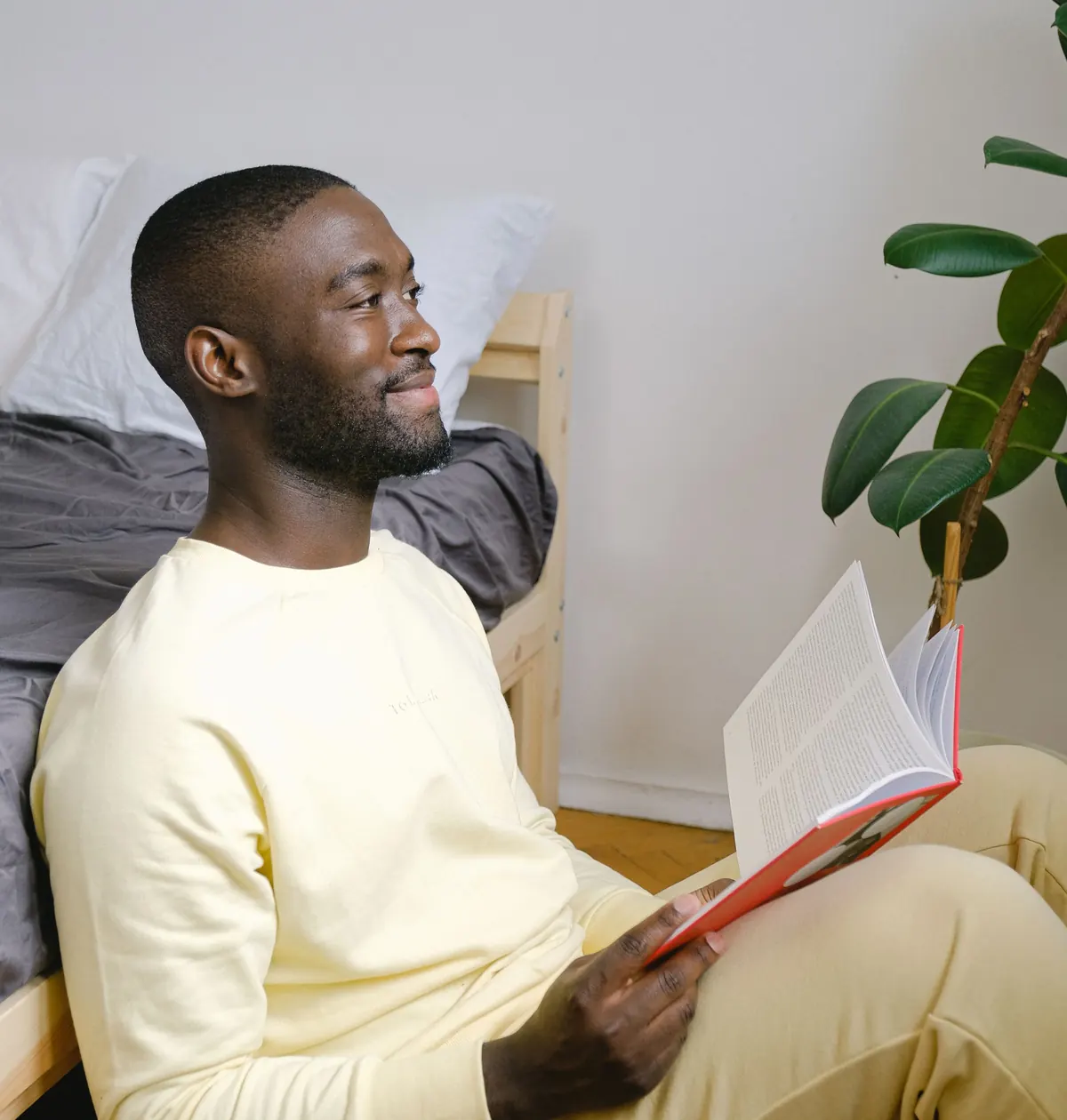 A man in a pale yellow sweater sits on the floor with a book open on his knee, smiling