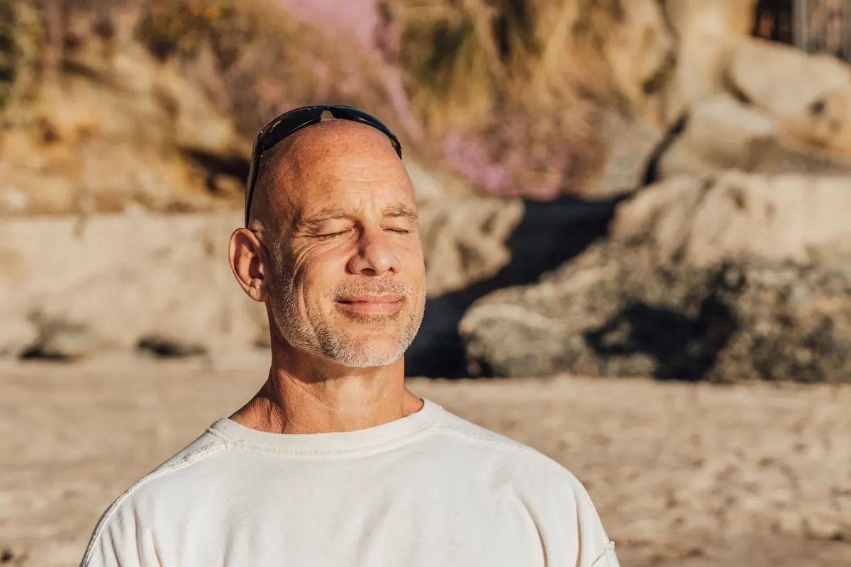 A smiling man sits in the sun on a sandy shore, eyes closed