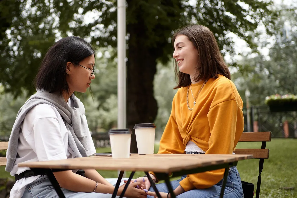 Two women talk at an outdoor cafe table, holding hands