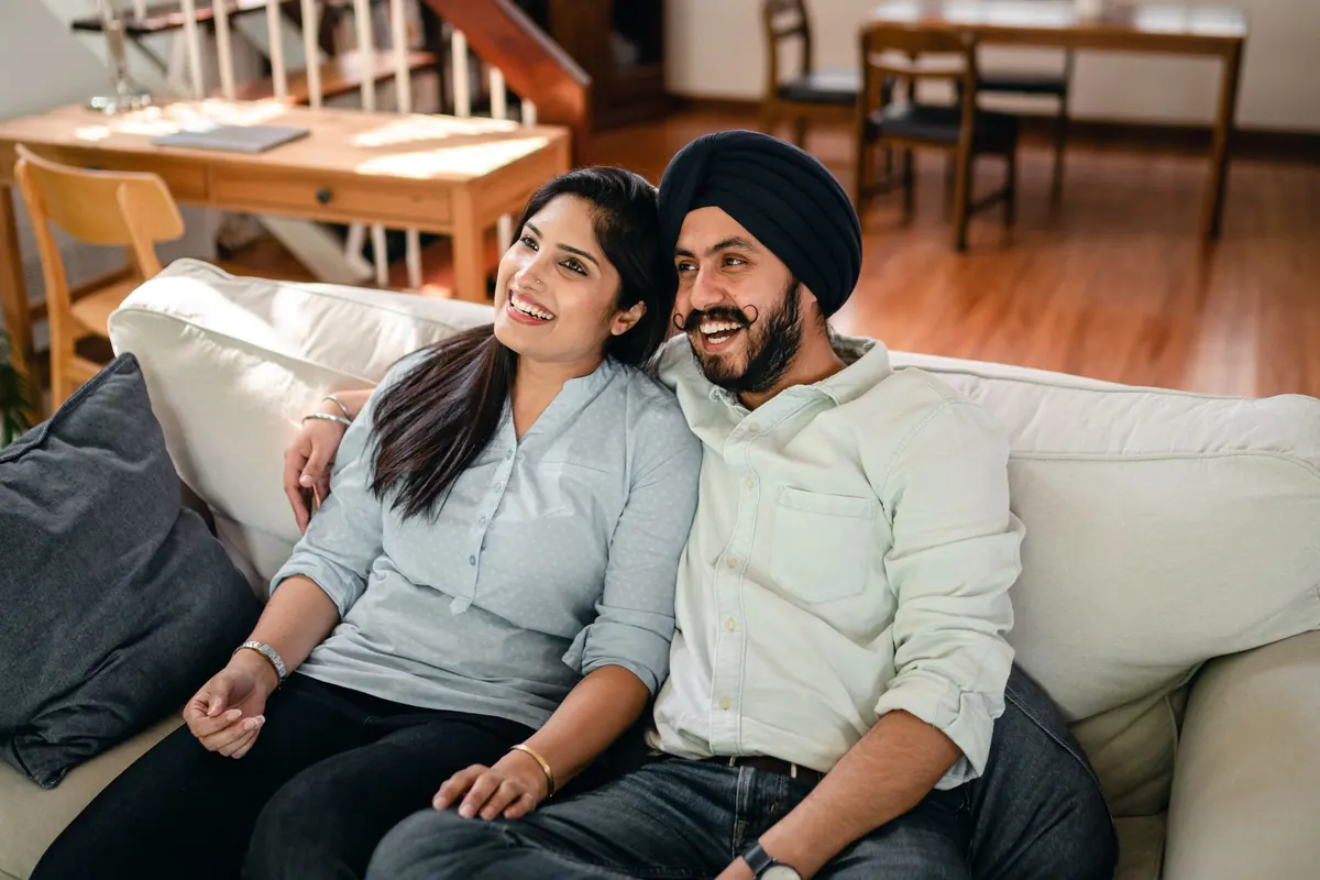 A couple sit close together on a sofa at home, laughing