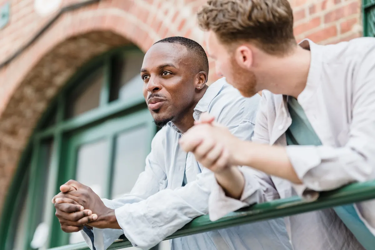 Two men lean on a railing outside a brick building, deep in conversation