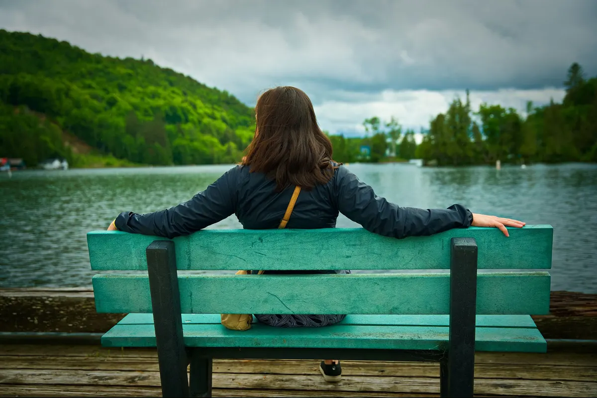 A woman sits on a bench by a lake, arms resting along its back