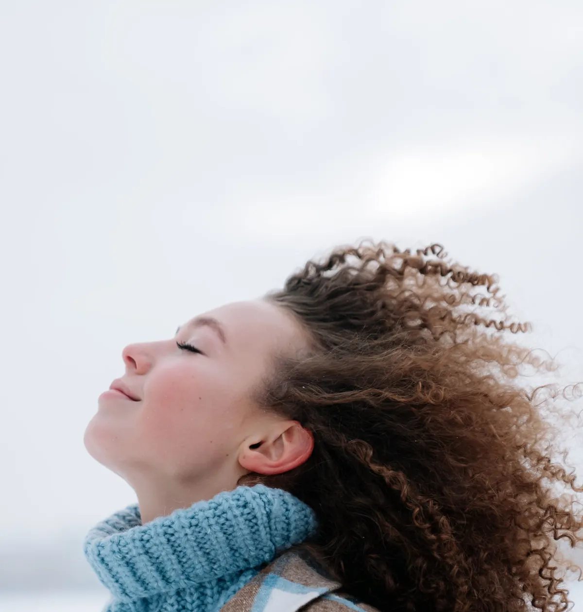 A woman with curly hair tilts her face to a pale winter sky, eyes closed, breathing in the cold air