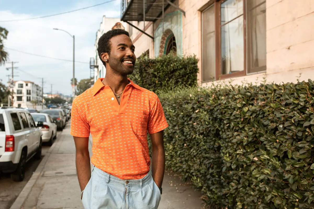 A smiling man in an orange shirt walks along a sunny city sidewalk lined with hedges