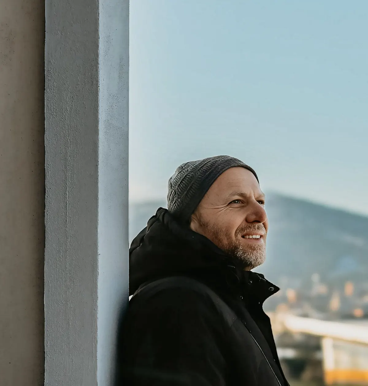 A man in his forties in a warm hat and jacket leans against a sunlit wall, smiling as he looks out over the hills