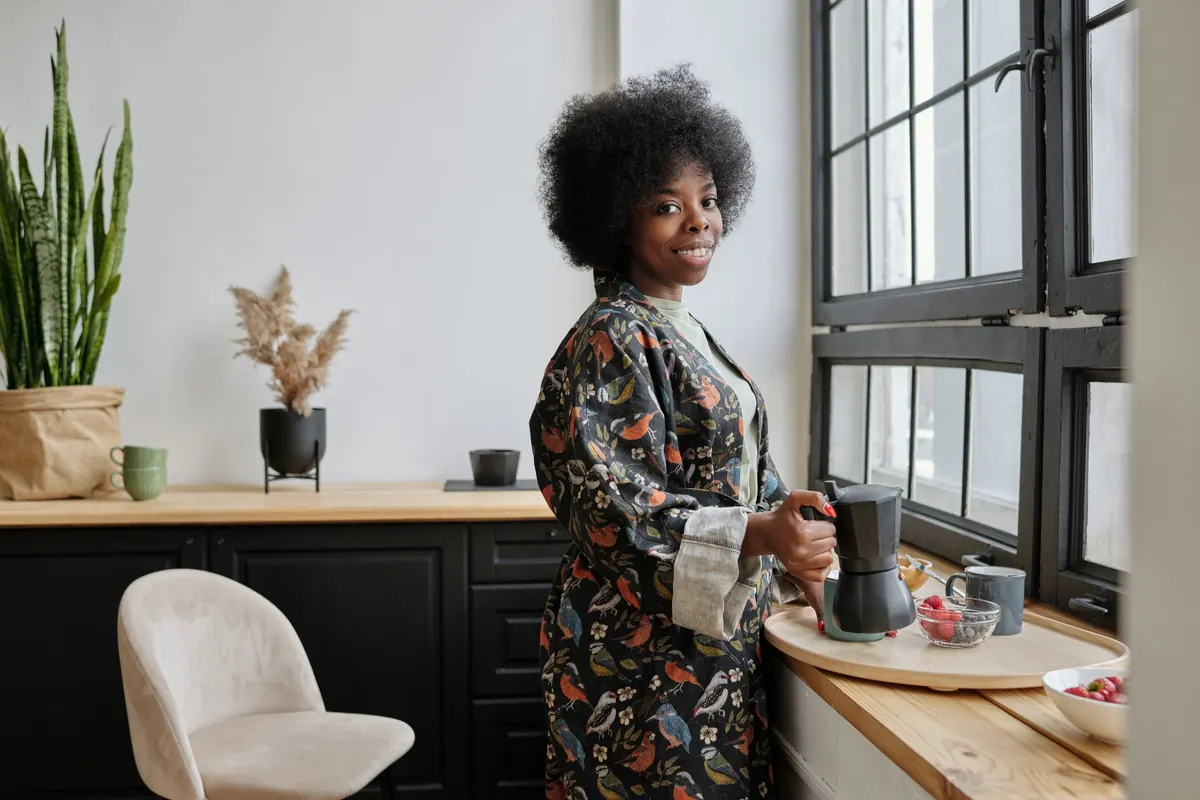 A smiling woman makes coffee by a sunny kitchen window