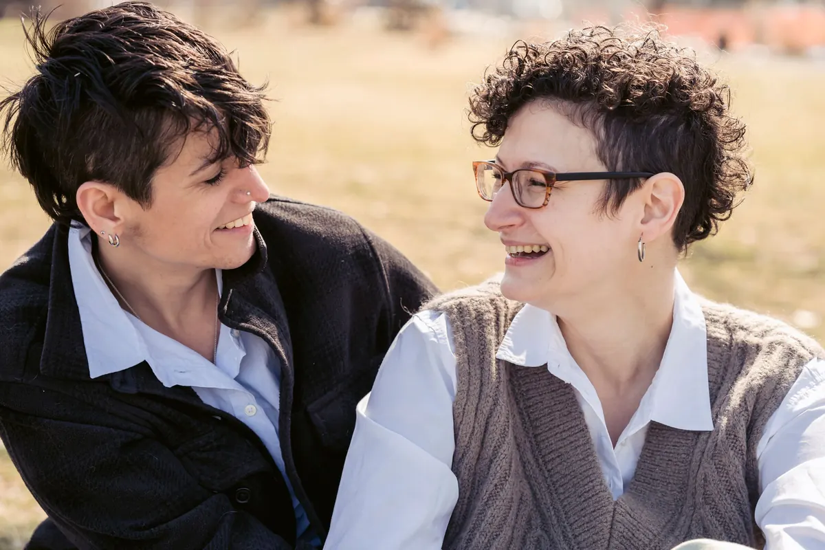 Two women laugh together, sitting close on the grass on a sunny day