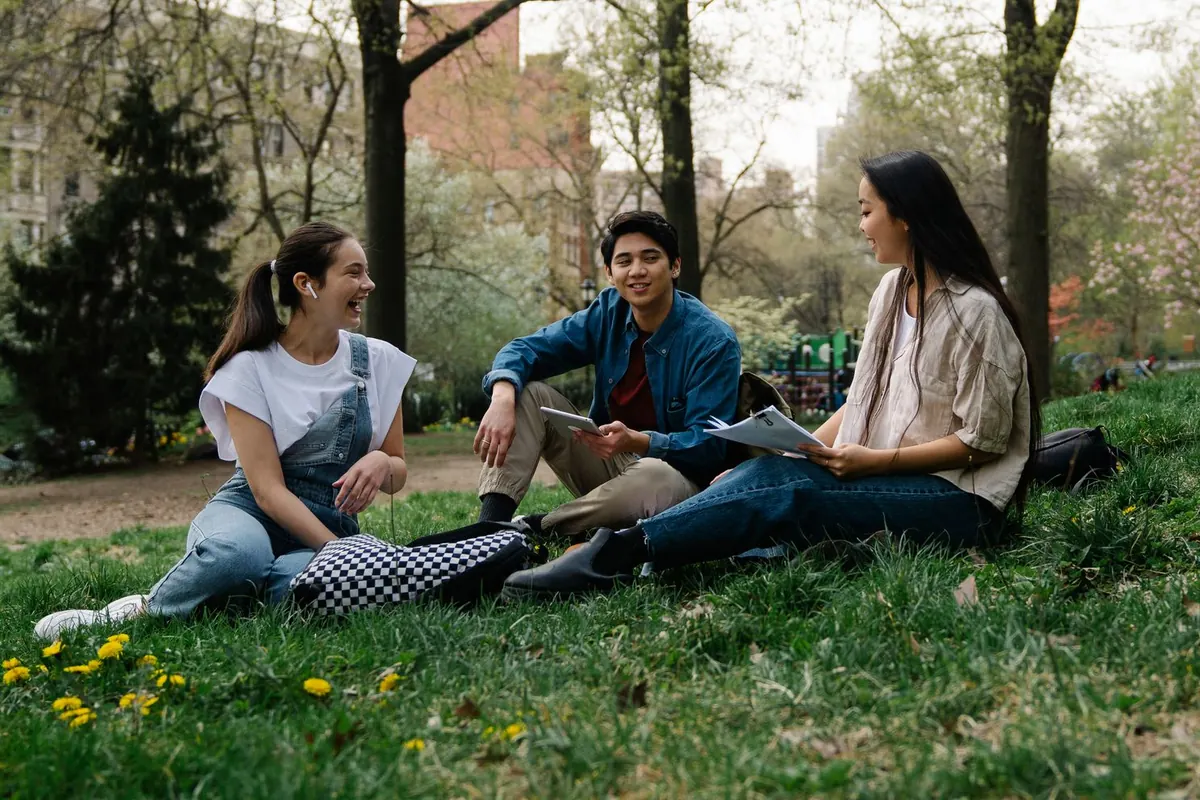 Three friends sit on the grass in a city park, talking and laughing