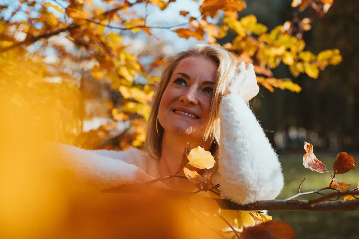 A smiling woman in a cream sweater resting her head on her arm among yellow autumn leaves